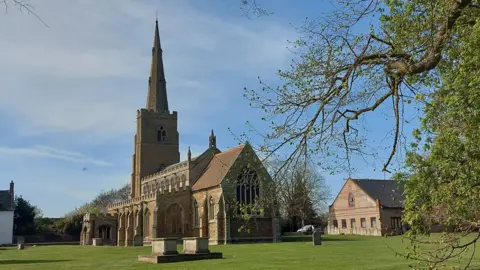 The Reverend Ruth Clay St Wendreda's Church in March - it has a tall spire. The churchyard has a few headstones on an otherwise open grassy expanse.