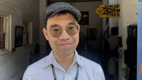 A man with a flat cap, round sunglasses and a pale shirt in front of a building with white washed walls.
