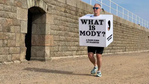 A man wearing a sugar cube costume which says "WHAT IS MODY" and "A RARE FORM OF DIABETES" on the front of it. He is wearing black cycle shorts. He is running on brown gravel. He is wearing sunglasses. Behind his is a brown wall. The sky is blue.