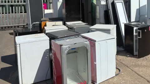 About 10 appliances, with around half of them white, are near each other outside. Railings are in the background on the left.