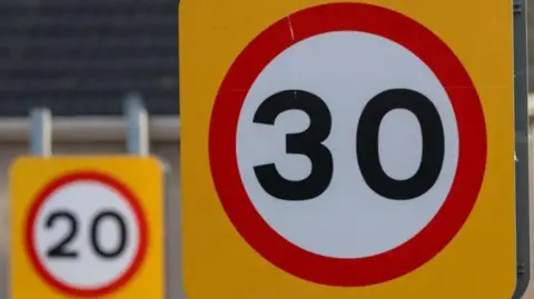 Two speed limit signs. They are square-shaped with an orange background. In the centre is a red circular band around a white centre with the numbers 20 and 30 inside in black.
