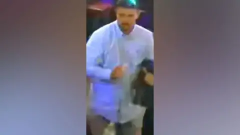 CCTV image showing a man with dark hair and short facial hair wearing a blue shirt and jeans.