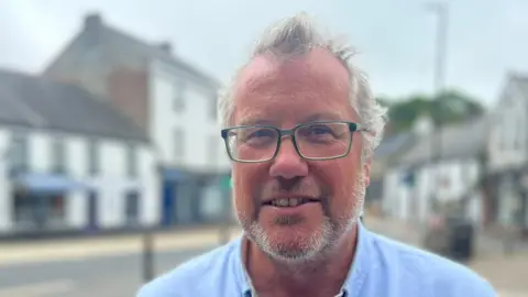 Councillor Mark Burnett smiles at the camera while stood in Camelford town centre, which is blurred behind him.