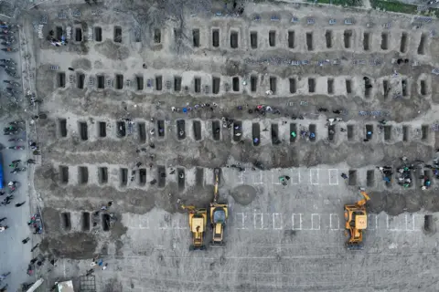 Iranian Press Center/AFP via Getty Images An aerial picture shows a series of graves dug on the ground with people around them. Yellow tractors are also in view