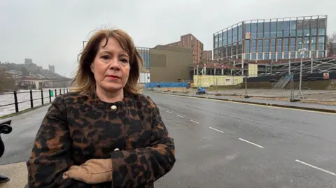 MP Mary Kelly Foy standing across the road from the almost finished Milburngate development. Foy is looking at the camera with a serious expression. She has shoulder-length brown hair and is wearing a leopard print-type coat with golden buttons. To the left is the river Wear and to her right is the development. It is made of a main building with window walls that looks finished. In front of it is heras fencing around the perimeter.