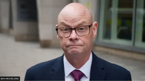 A bald man with glasses, wearing a dark suit and tie, is pictured on a pavement, with a building behind him