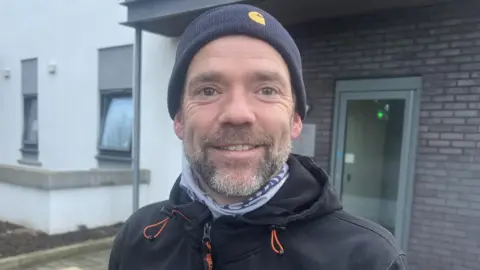 Andrew McCabe is wearing a black jacket, black hat and has a beard. 