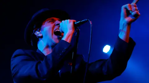 A close-up of Paul Smith on stage. He is wearing a wide-brimmed hat and a dark suit jacket. The camera is underneath him and he is pointing to the right and singing into a microphone. The light is blue and casting him partly in shadow.