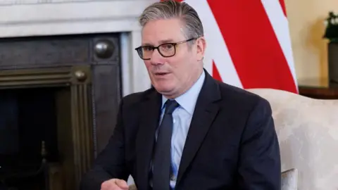 Prime Minister Sir Keir Starmer sat in a white chair in front of a Union flag. He is wearing dark-rimmed glasses, a dark-coloured suit jacket, a light blue shirt and a tie.