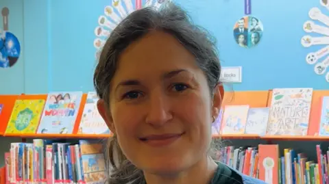 Jacob Ottaway/BBC Lizzie Edwards smiles at the camera inside a library. Books can be seen stacked on a shelf behind her. She has brown hair that is tied back behind her head.