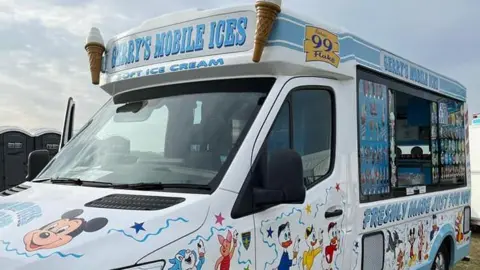 The outside of an ice cream van, it is white, with Disney characters painted in it, the window is open, with pictures of the ice creams for sale on the side. A door is open to the van and there are portable toilets to the left. 