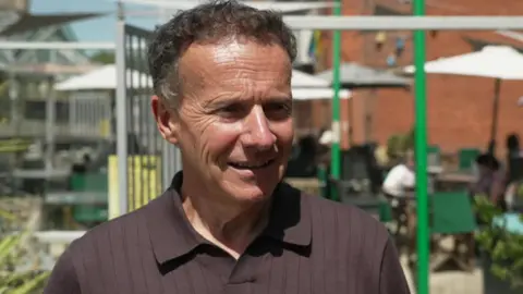 A man standing outdoors, facing slightly to the right of the camera and wearing a dark, short‑sleeved polo shirt. There is an outdoor seating area with tables, chairs and large white parasols behind metal railings in the background. Green plants in containers and a brick building are further back. The scene is well lit, as it is a sunny day.
