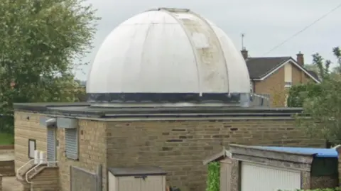 LDRS A small observatory building made of light brown brick with a flat roof and a large white dome on top, which appears weathered and slightly stained.