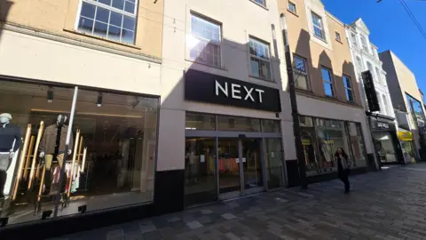 A high street store with large glass windows and a central double door opening. Above the doors in a large black rectangular sign with "NEXT" written in white. It is a sunny day but the lower part of the store is covered by the shadows of buildings opposite.