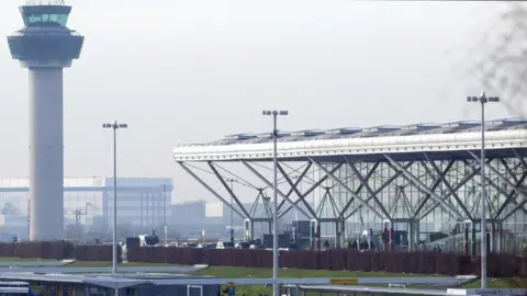 PA Media London Stansted Airport on a cloudy day. The airport control tower is to the left and the main terminal buiding, which has a glass facade and a white roof supported by grey pillars.