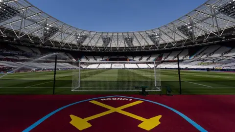A wide-angle view from behind a football goal net looking into the empty London Stadium under a clear blue sky, featuring the West Ham United crossed hammers logo painted in yellow on the ground in the foreground.