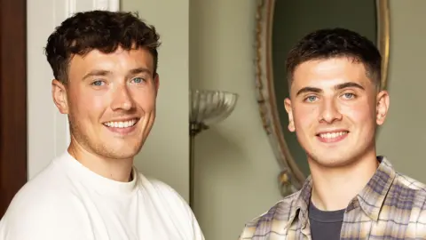 Jordan and Cian are pictured together inside a house. Jordan (on the left) has dark curly hair and is wearing a white t-shirt. Cian (on the right) has short, dark hair and is wearing a dark grey t-shirt and a checkered shirt over the top.