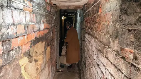 Emma and Daisy are at the end of a narrow brick tunnel talking to a man who is sitting on the ground