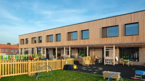 Morgan Sindall/Buntingford First School The school's exterior, at a slight angle, showing a two-storey building. The ground storey is made of brick and glass, but the top floor is entirely clad in wood, and has large windows which have wooden slats partly covering them. Outside is a play area with black flooring and grass, and colourful wood fences.