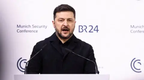 Reuters Volodymyr Zelenskiy in a dark coat stands at a podium with two microphones, speaking in front of a white backdrop displaying the Munich Security Conference and BR24 logos.