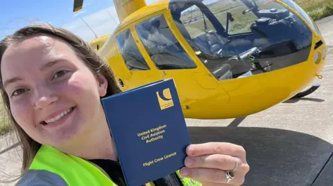 OLIVIA WATSON Olivia smiling while holing up her licence, which is in a blue cover, with the helicopter in the background.