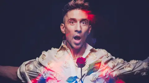 An artistic image of comedian Russell Kane - a man with short dark hair, wearing a white shirt with white flowers on it. A rose is also in the foreground. His mouth is wide open and his arms are open.