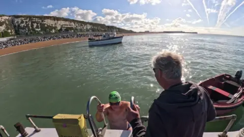 Grimsby and Cleethorpes Water Rats Open Swimming club Man wearing green swimming hat and black trunks climbing into a boat. There is a man with grey hair wearing a black hoodie standing on the boat. There's a red boat near them. The water is a green/blue. The sky is blue and sunny with white clouds. There are cliffs in the background and a white boat.