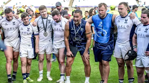 Pat Lam (fourth from left) stands with his hands on his knees surrounded by four players to his right, and a coach and two more players to his left, as he leads a post-match huddle on the field following their win against Bulls in Pretoria