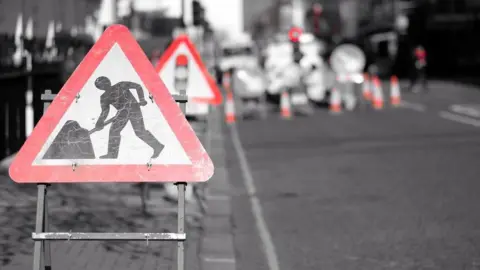 Road repair signs highlighted in red against the black and white of a road. The signs indicate roadworks and that a traffic light system is in place