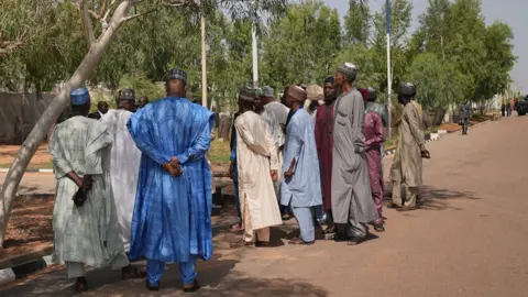 Un groupe d'hommes, vêtus de tenues nigérianes du nord, se tiennent à l'extérieur de la maison de Muhammadu Buhari. Il y a une rangée d'arbres verts derrière eux.