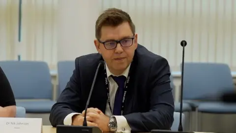 Dr Johnson wearing a black suit sitting in front of a microphone 