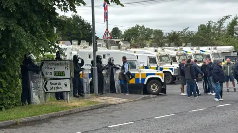 PA Five white, blue and yellow police vans block an entrance. A group of men are standing near the vans. Five police men wearing riot gear are standing next to the police vans.