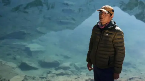 Peruvian farmer Saul Luciano Lliuya poses for a photo in front of Lake Palcacocha. He is wearing a baseball cap and behind him the clear waters of the lake can be seen. 
