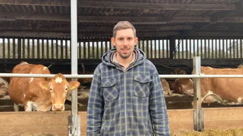 A young adult man who is wearing a navy chequered jacket with a collared shirt underneath. In the background there are cows in a barn one is looking at the camera while the rest are sitting down.