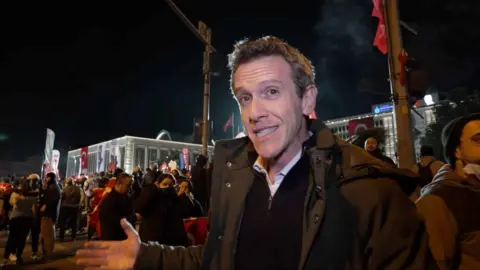 BBC correspondent Mark Lowen stands in front of a crowd of protesters in Istanbul at night.