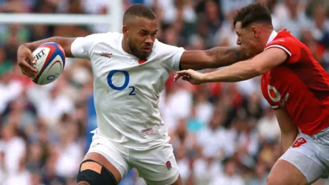 Reuters A male rugby player wearing a white England kit, holds the rugby ball in his right hand while holding off another player, wearing a red and white Wales kit, with his left hand.