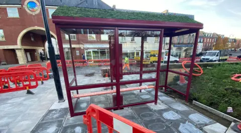 Wiltshire Council A new bus shelter in the town on freshly-laid paving slabs and surrounded by red barrier gates and a workman in a high vis jacket in the background