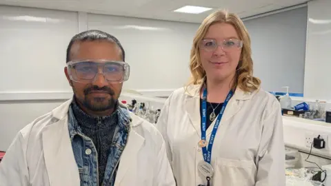 A man stands on the left wearing a white lab coat over a denim jacket with safety goggles over his glasses. He has short dark hair and dark facial hair. To his right, a woman stands wearing a white lab coat done up with a blue staff lanyard and necklace on. She has shoulder length curled blonde hair, behind then is a lab desk.
