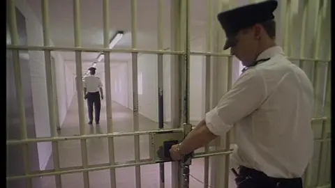 A prison officer locks a pair of metal gates at the end of a corridor. Further away is another prison officer. They are wearing white shirts, black trousers and have peaked black caps