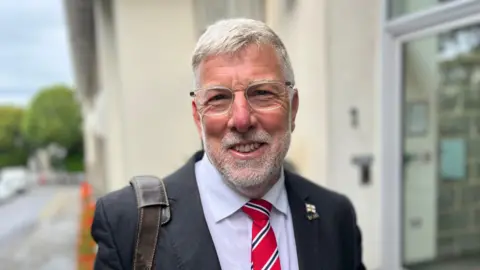 Deputy Adrian Gabriel - A man with grey hair, clear rimmed glasses and a grey beard. He's wearing a black suit, white shirt and a red and blue striped tie. 