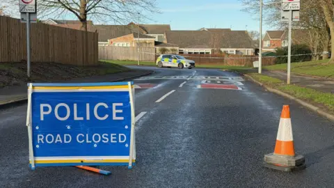 A blue sign reading POLICE ROAD CLOSED next to an orange traffic cone on Carr Lane in Spennymoor. A police car is parked further down the road. Part of the pavement behind the car is fenced off with a tape. 