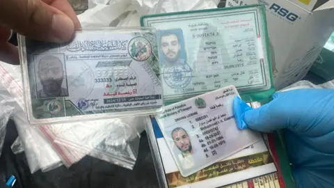 IDF A selection of identification documents are held by two hands, one of which wears a plastic glove, with evidence bags in the background.