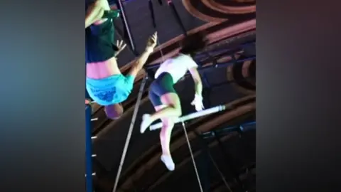 Two trapeze artists practising. One is in mid-air about to grab her trapeze, while a man is hanging on his trapeze by his legs, having just released her.