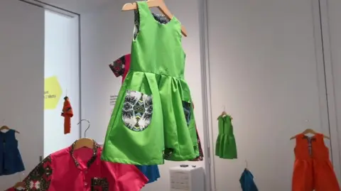 Exhibit of brightly-coloured children's dresses on clothes hangers hanging from the ceiling, set against white walls