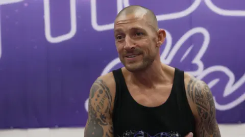 Elmo is standing in front of a purple banner, smiling. He is wearing a tank top and has tattooed arms and a shaved head.