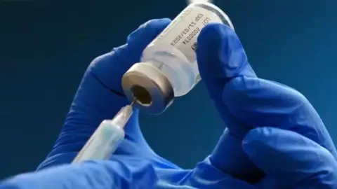 A pair of hands in blue rubber gloves pushing the needle of a syringe into a vial of vaccine.