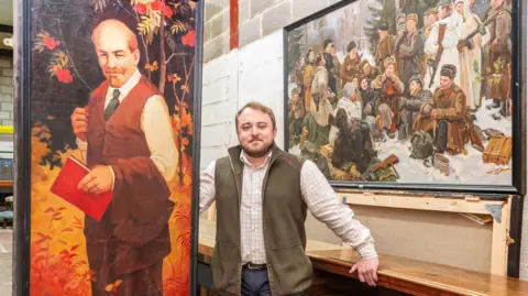 Wotton Auction Rooms Joseph Trinder looks into the camera, wearing a check shirt and green gilet, while standing beside the tall painting of Lenin, which is quite a lot larger than him, to show its scale.