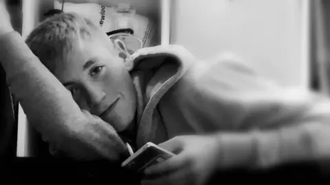 PA Media A black and white photo of Jack, who looks straight at the camera, and is lying down, leaning on one arm as he holds his phone in his other hand. He has short hair and is clean shaven, and is wearing a hoodie. 