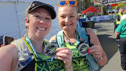Kate Walford and Laura Coleman-Day are holding up Guernsey Marathon finishing medals and wearing running gear and sunglasses. They are grinning broadly. 