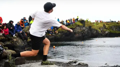 Shows a man standing on a rocky ledge and throwing a stone across the water while spectators watch from the opposite shore.